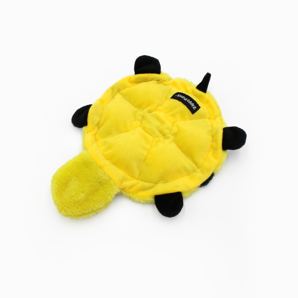 A plush, yellow turtle-shaped dog toy with a black label and black feet and head is shown on a plain white background.