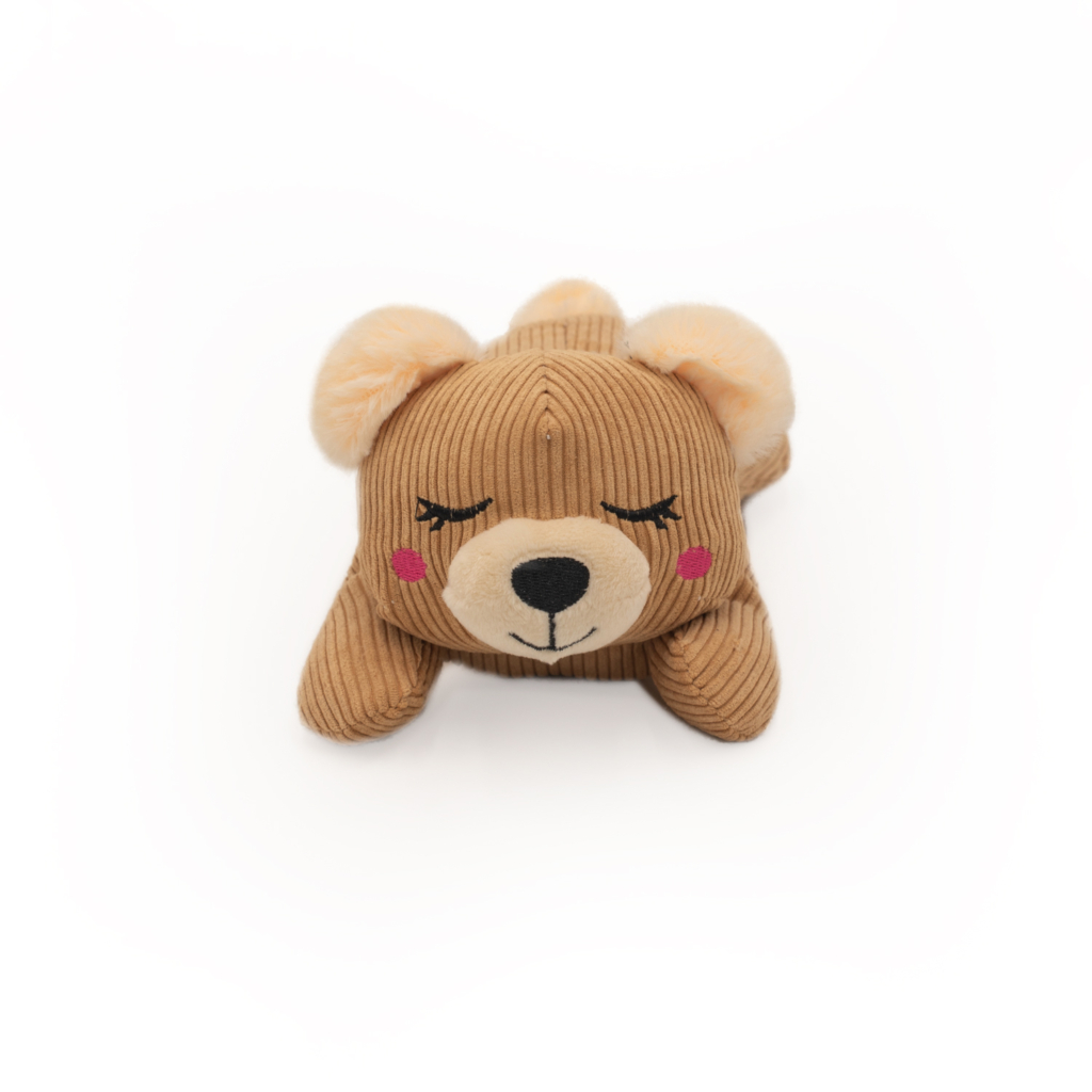A brown, plush Snooziez with Shhhqueaker - Bear with closed eyes, pink blush on its cheeks, and a neutral expression, lying on its stomach. The background is plain white.