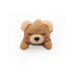 A brown, plush Snooziez with Shhhqueaker - Bear with closed eyes, pink blush on its cheeks, and a neutral expression, lying on its stomach. The background is plain white.
