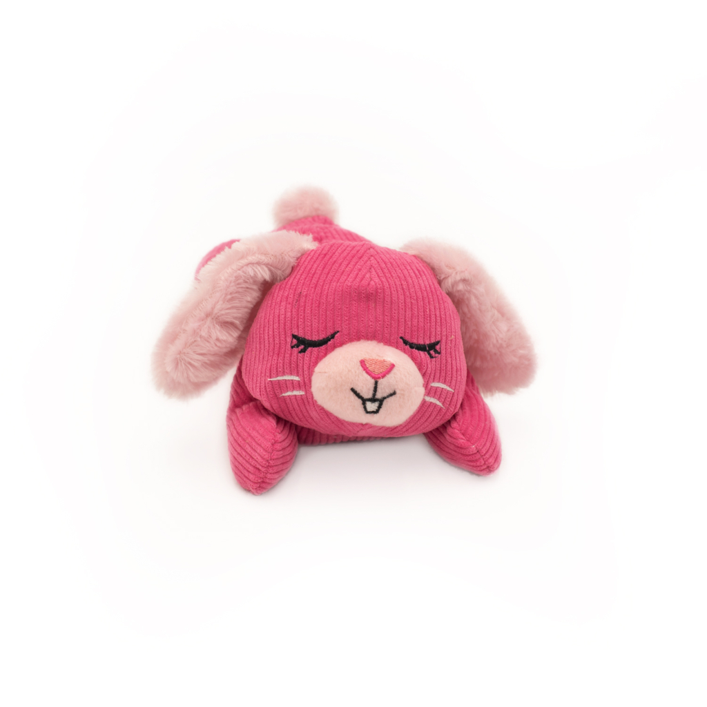 A small, pink Snooziez with Shhhqueaker - Bunny plush toy resembling a bunny with closed eyes, pink ears, and a smiling face is lying on a white surface.