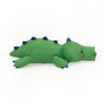 A green, plush Snooziez with Shhhqueaker - Alligator lying on a white background with dark blue spikes along its back and a red blush spot on its cheek.