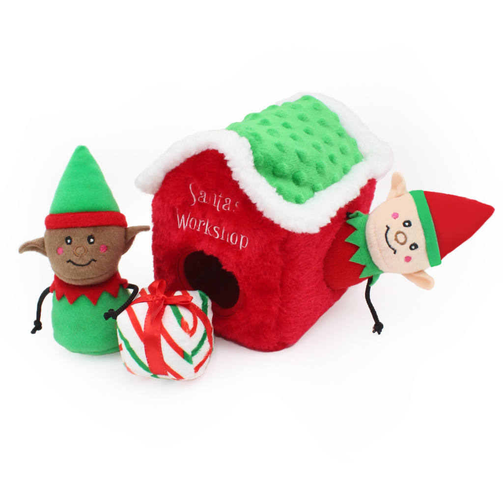 Plush elf toys and a candy cane-striped plush present are placed around a red Holiday Burrow® - Santa's Workshop. One elf is inside the house, and the other stands outside.