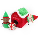 Plush elf toys and a candy cane-striped plush present are placed around a red Holiday Burrow® - Santa's Workshop. One elf is inside the house, and the other stands outside.