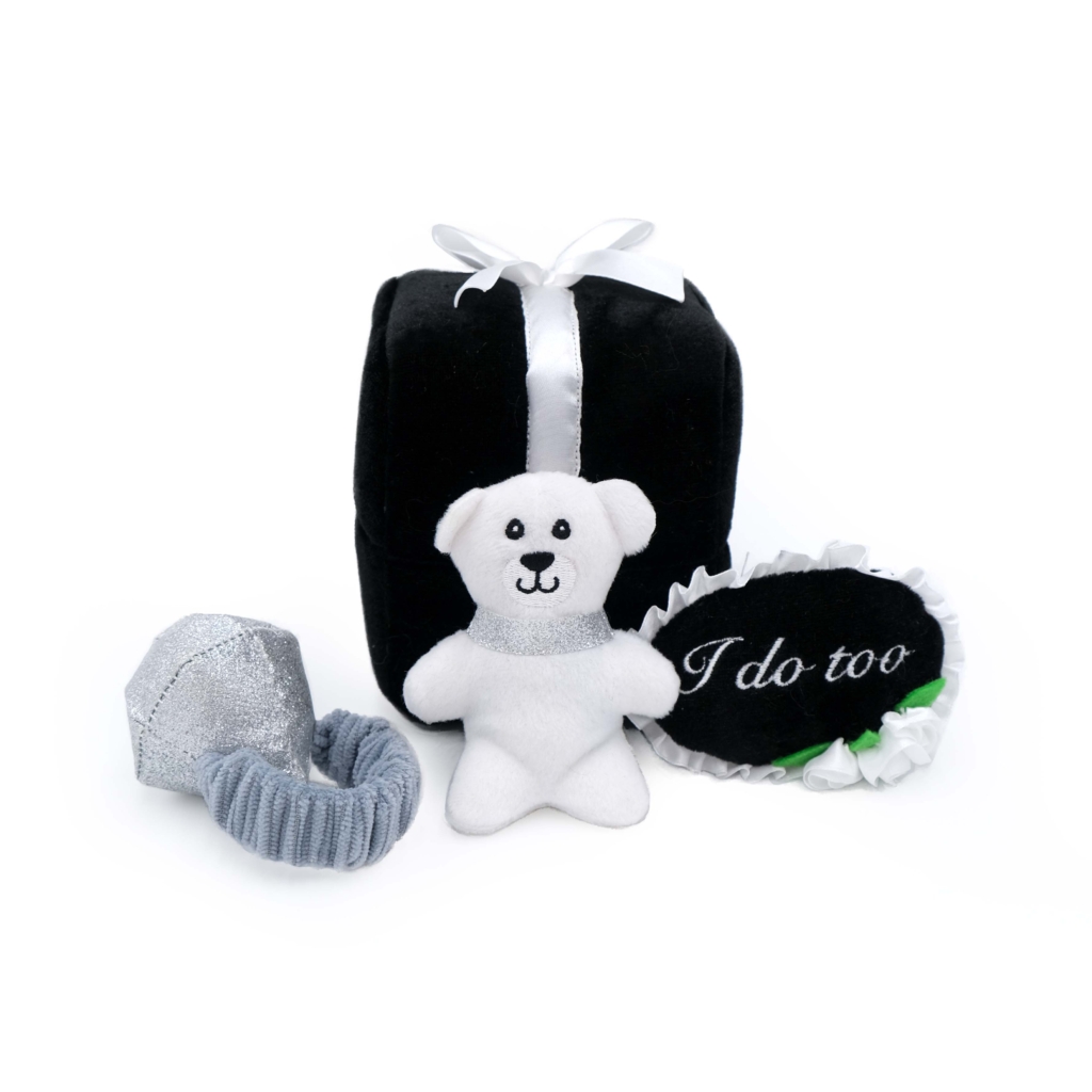 Plush white teddy bear with a gray scarf sitting next to a black gift box with a white ribbon, a Zippy Burrow® - Wedding Ring Box, and a black oval pillow with 