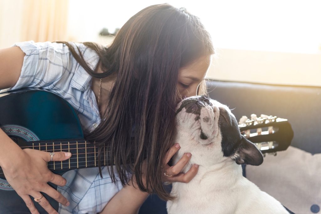 Young latin woman plays the guitar and kisses her dog (french bulldog)
