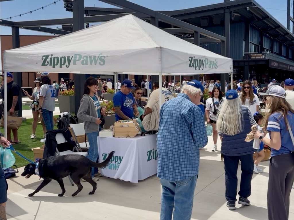 People gather around a ZippyPaws booth outdoors. A black dog on a leash walks by, and several people wear baseball caps and casual clothing. The booth is under a white canopy.