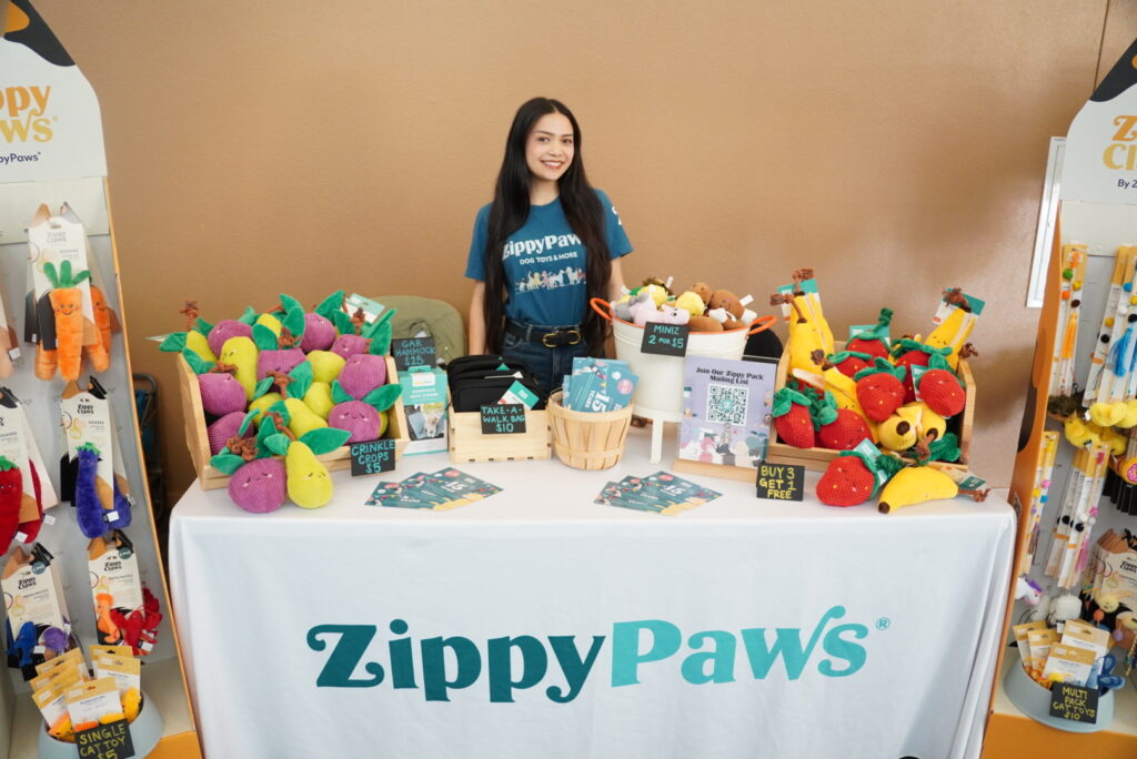 A woman stands behind a ZippyPaws display table featuring colorful pet toys, promotional flyers, and signs indicating sales and offers at a pet products event.