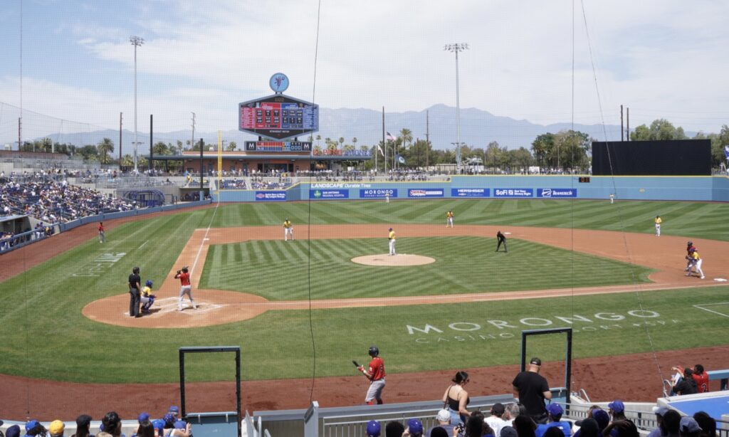 A baseball game in progress at a stadium, with players on the field, spectators in the stands, and mountains visible in the background.