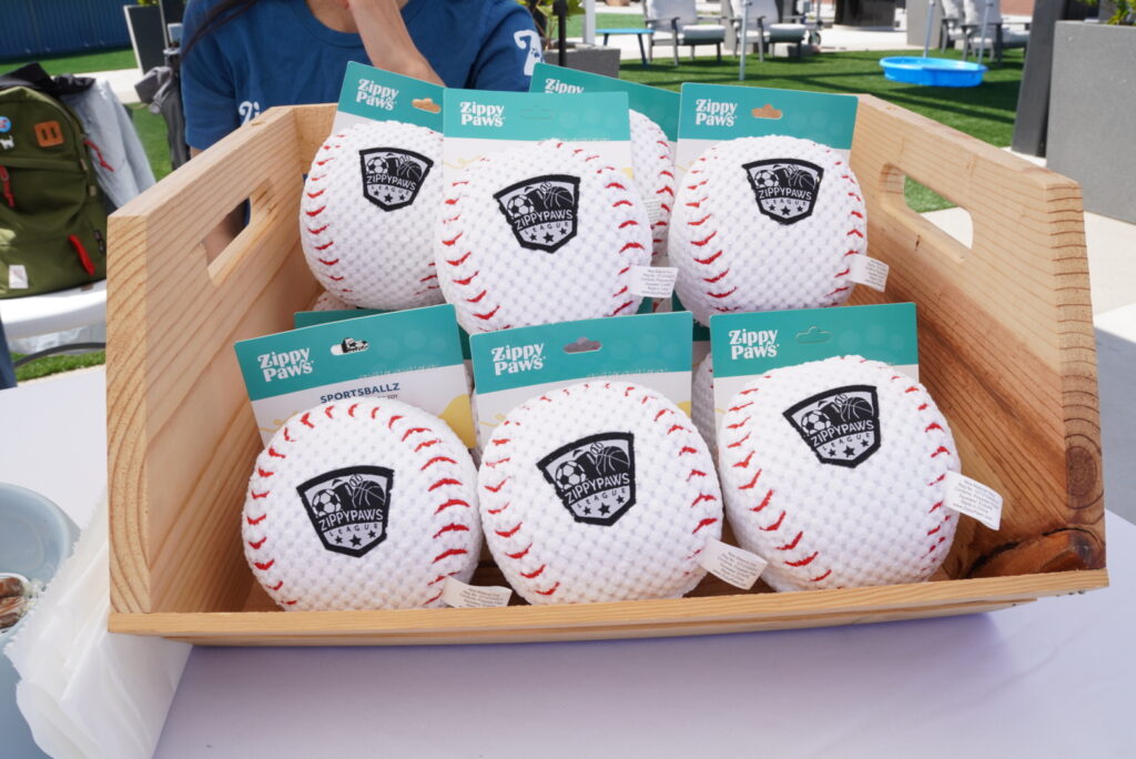 A wooden crate holds six ZippyPaws plush baseball dog toys, each with a white surface, red stitching, and a black paw print logo.