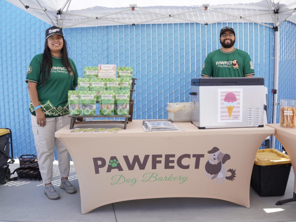 Two people stand behind a booth labeled "Pawfect Dog Barkery," displaying dog treats and snacks under a canopy at an outdoor event.