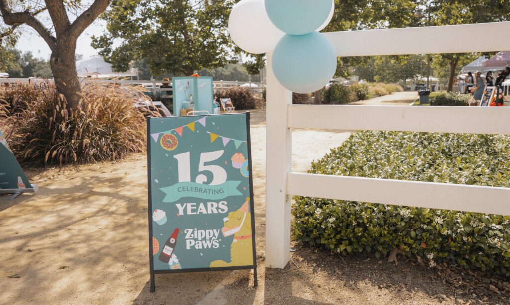 A sign reads "15 Years Celebrating Zippy Paws" with festive graphics, set outside on a dirt path near a white fence and greenery, with balloons attached to the fence.