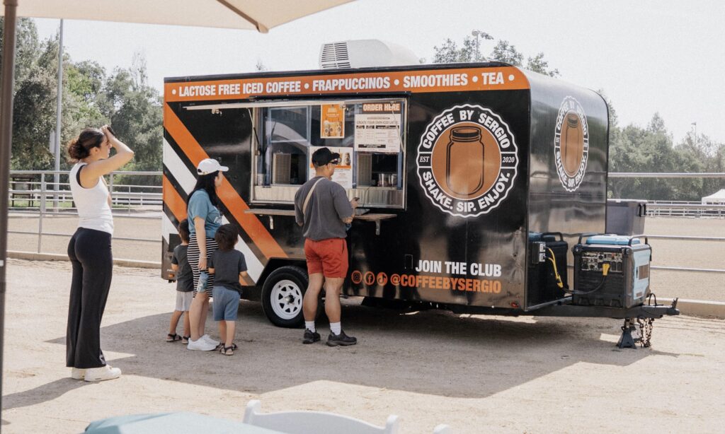 People stand in line at a black coffee truck offering lactose-free iced coffee, frappuccinos, smoothies, and tea in an outdoor setting.
