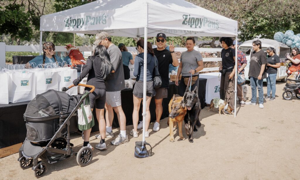 People gather under a white "Zippy Paws" tent at an outdoor event. Some are holding dogs on leashes, while others stand nearby. Gift bags and balloons are visible in the background.