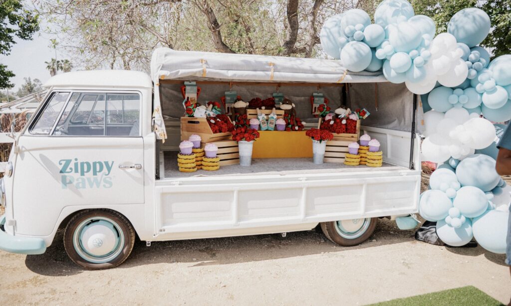 A white van with "Zippy Paws" branding displays plush dog toys arranged in crates. Blue and white balloons decorate the side of the van, which is parked outdoors.