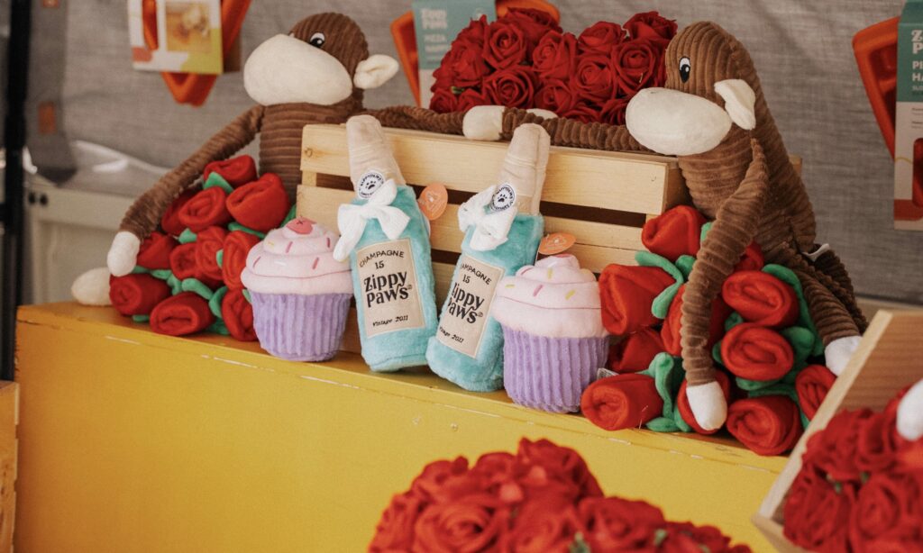 Plush monkey toys, red rose bouquets, and cupcake and bottle-shaped plushies displayed on a yellow counter with a wooden crate in the background.