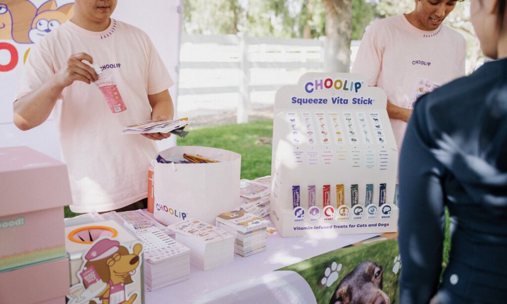 Two people at an outdoor booth display and distribute CHOOLIP Squeeze Vita Stick pet treats, with product samples and informational materials on a table.