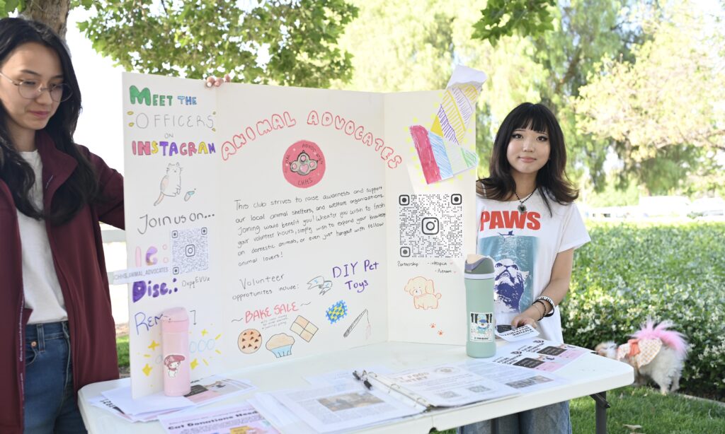 Two young women stand at an outdoor table displaying an "Animal Advocates" trifold board with information, drawings, QR codes, and sign-up sheets for a volunteer club.