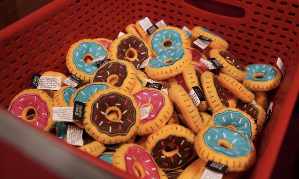 A red plastic basket filled with plush toy donuts in blue, pink, and brown colors, each with embroidered sprinkles and tags attached.