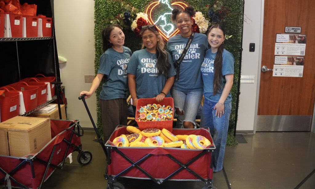 Four women wearing matching blue shirts stand together indoors, smiling behind a cart filled with colorful donut-shaped plush toys. Shelves and bins are visible in the background.