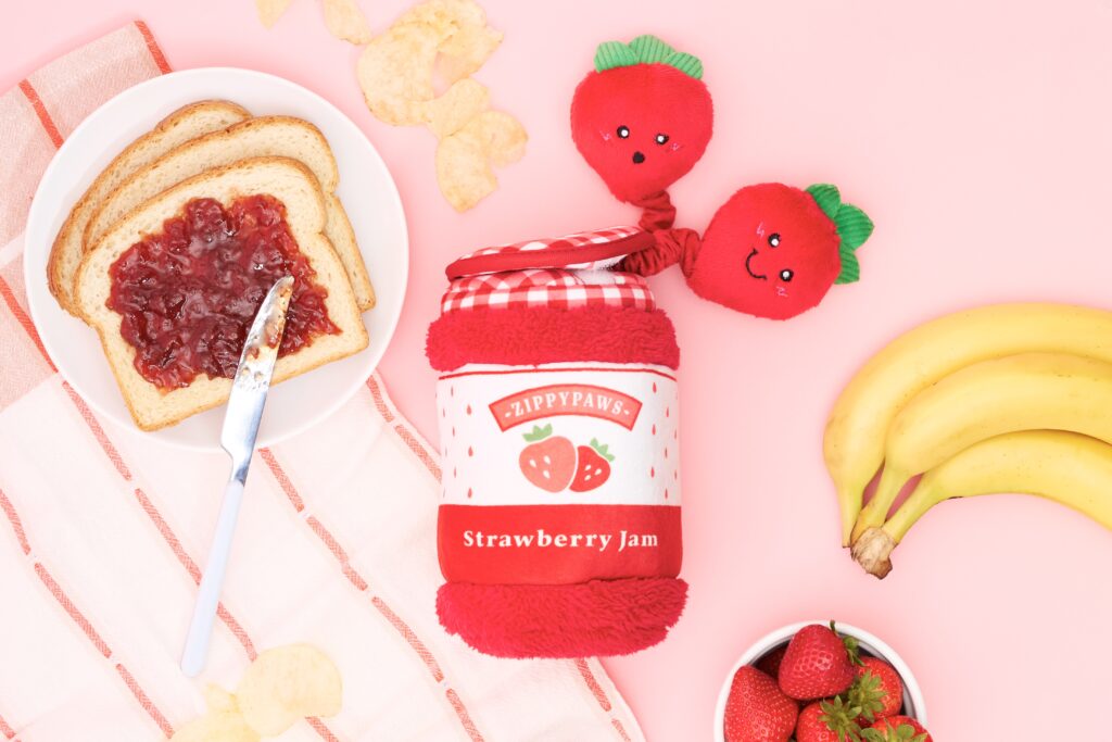 A jar of strawberry jam with two plush strawberries, sliced bread with jam, a butter knife, bananas, potato chips, and fresh strawberries on a pink surface.