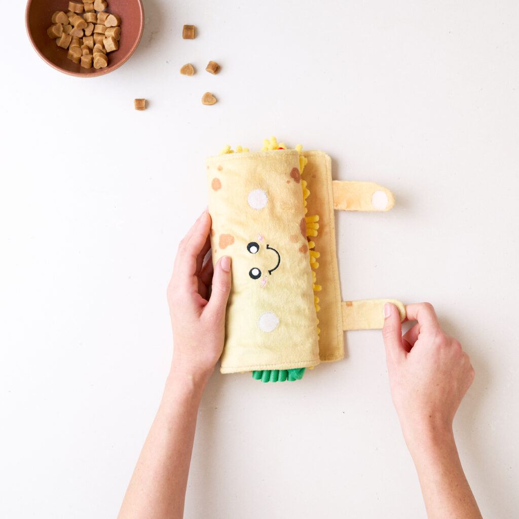 Hands holding a plush toy shaped like a smiling burrito next to a small bowl of brown treats on a white surface.