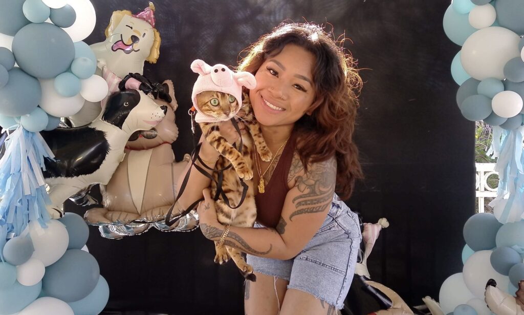 A woman with tattoos smiles while holding a cat wearing a pink hat, surrounded by blue and white balloons and animal-shaped decorations.