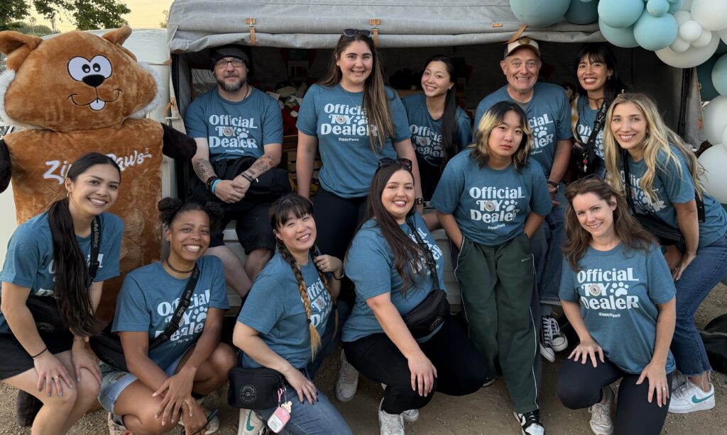 Group of people wearing matching "Official Toy Dealer" shirts pose outdoors in front of a tent, with a person in a brown mascot costume and blue balloons in the background.