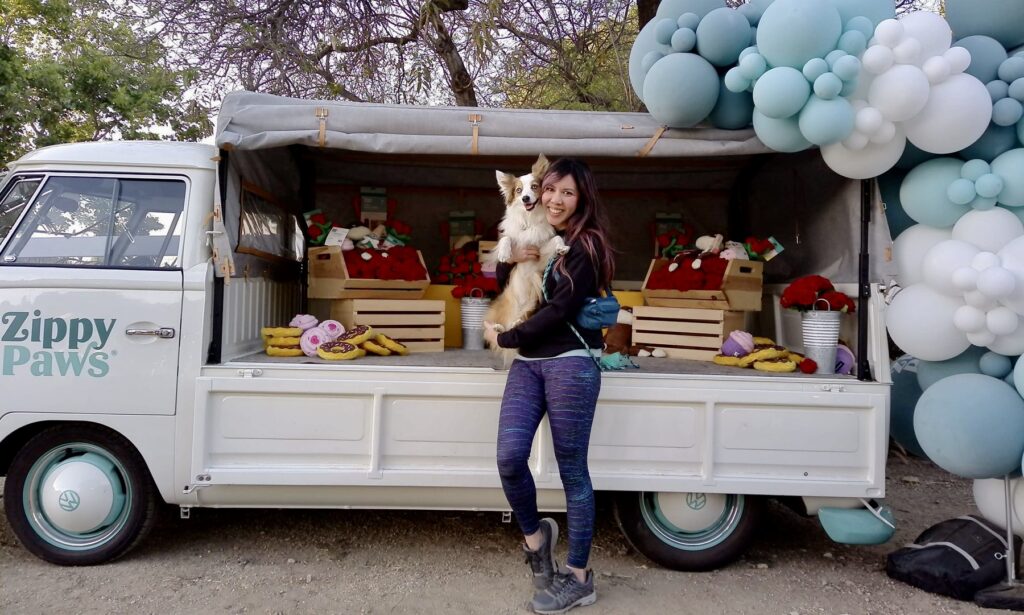 A woman stands in front of a decorated van holding a small dog, with dog toys and gift boxes displayed in the van, and blue and white balloons overhead.
