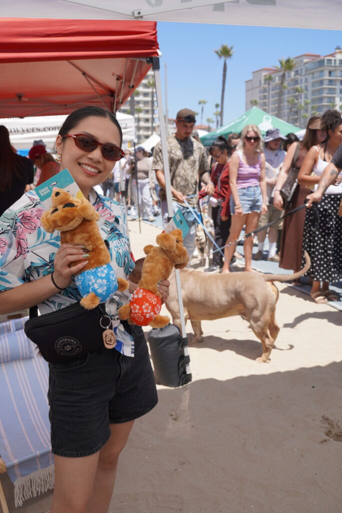 A person in sunglasses and a floral shirt holds two plush kangaroo toys at a beach event, with people and a dog in the background under tents.