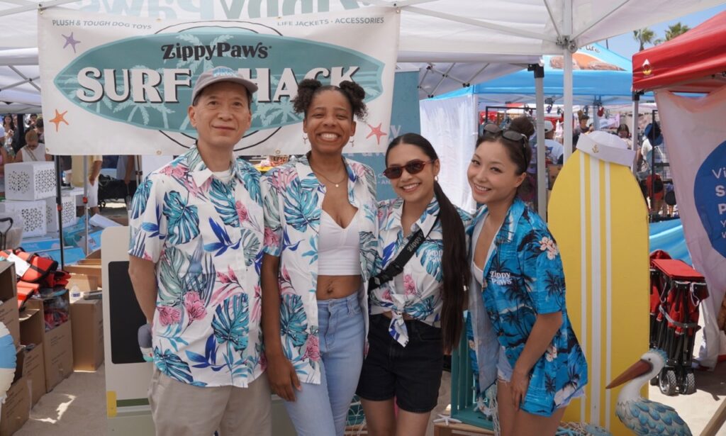 Four people wearing matching blue floral shirts pose and smile in front of a "ZippyPaws Surf Shack" booth at an outdoor event or festival.