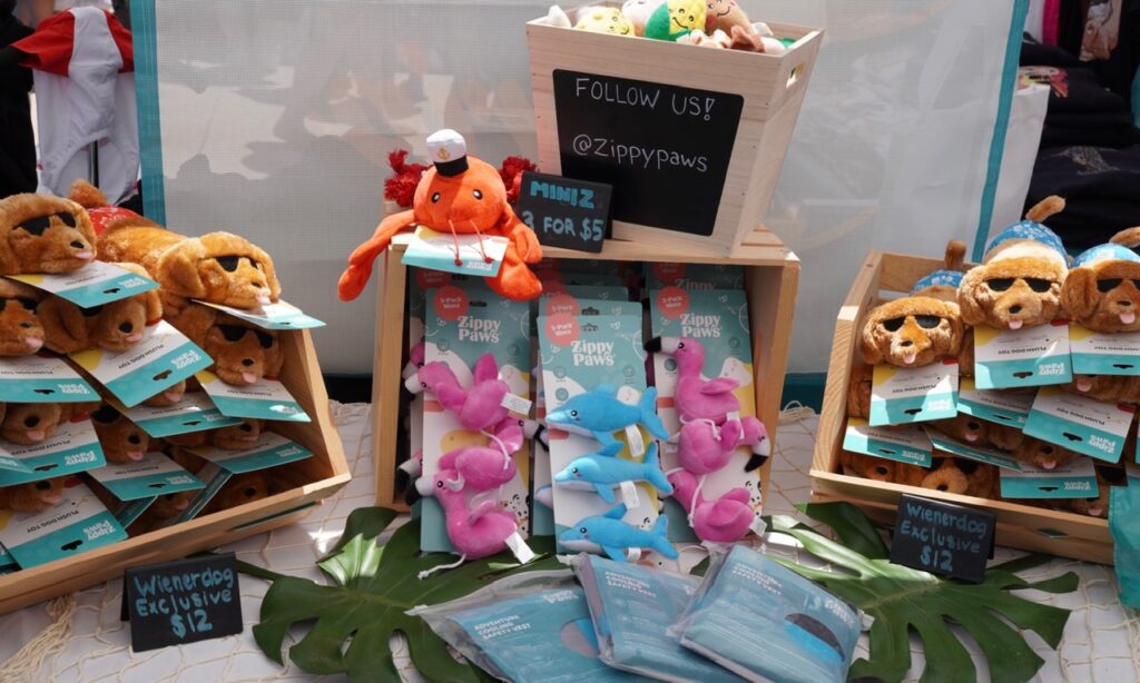 Display of plush dog toys, including wiener dogs, sharks, and octopuses, arranged in wooden crates with a sign promoting @ZippyPaws and prices labeled.