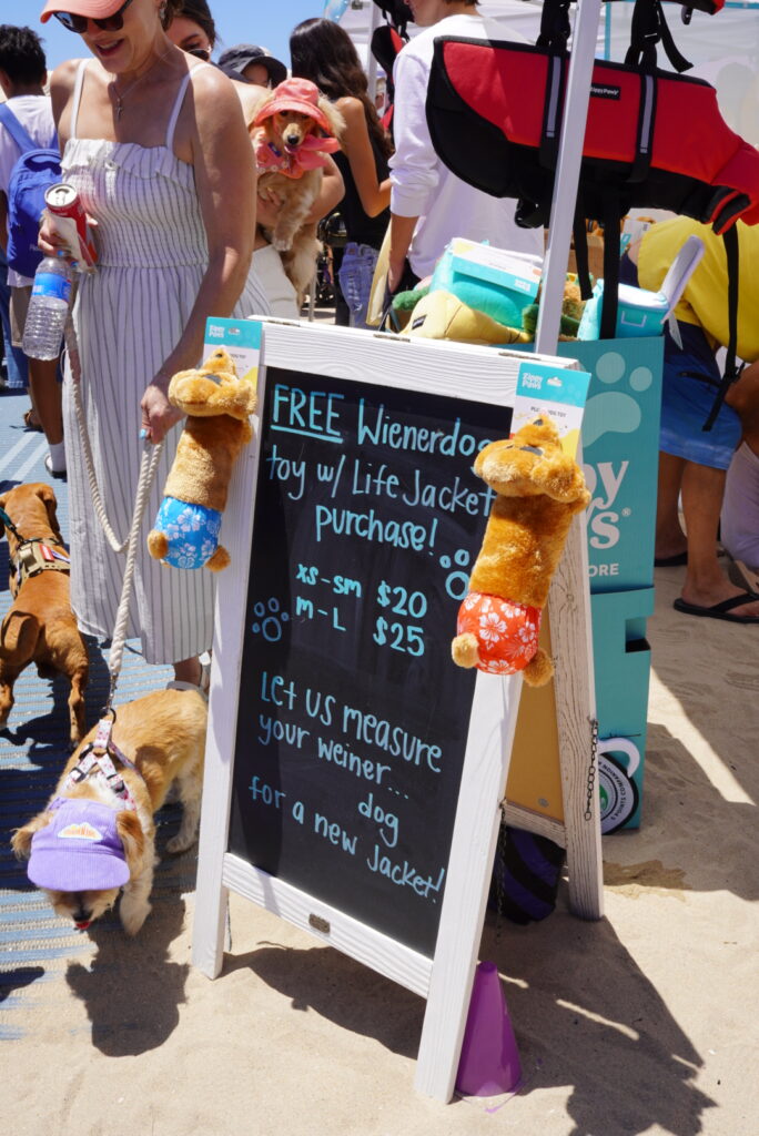 A chalkboard sign at an outdoor event advertises free dog toys with life jacket purchases, surrounded by people and dogs on leashes.