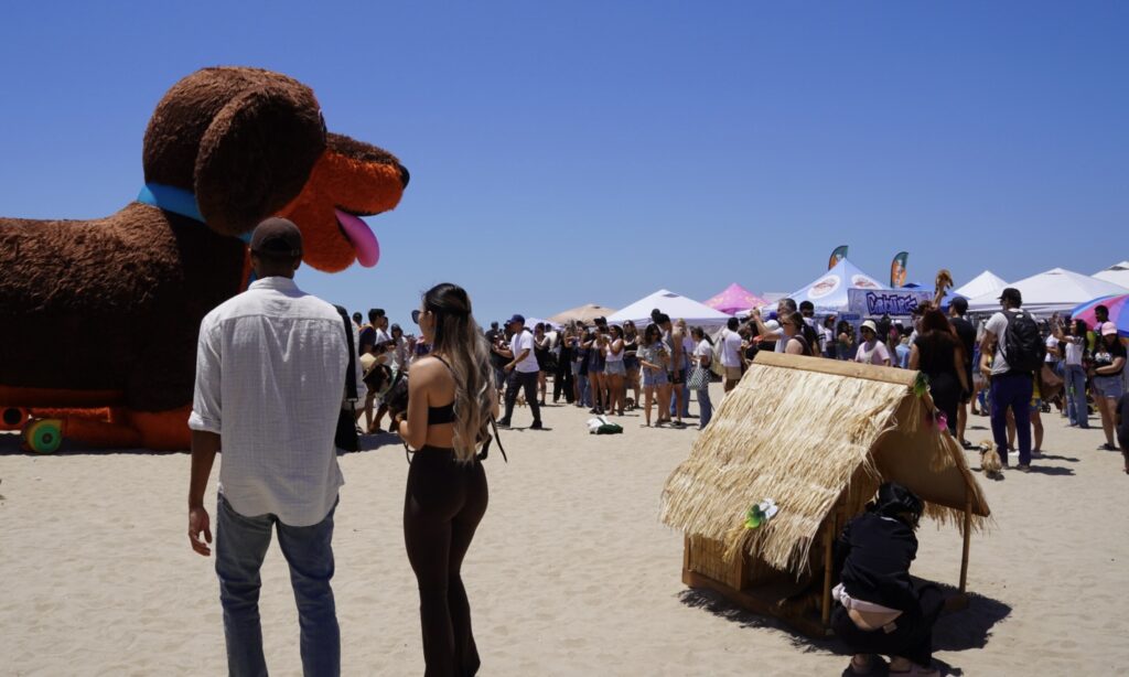 People gather on a sandy beach near a large inflatable dog and a small straw hut, with tents and a clear blue sky in the background.