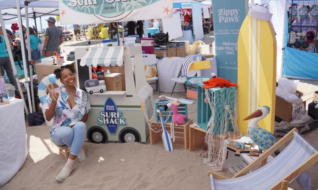 A woman poses and smiles in front of a beach-themed vendor booth featuring surfboards, a pelican statue, and a “Surf Shack” cart at an outdoor event.