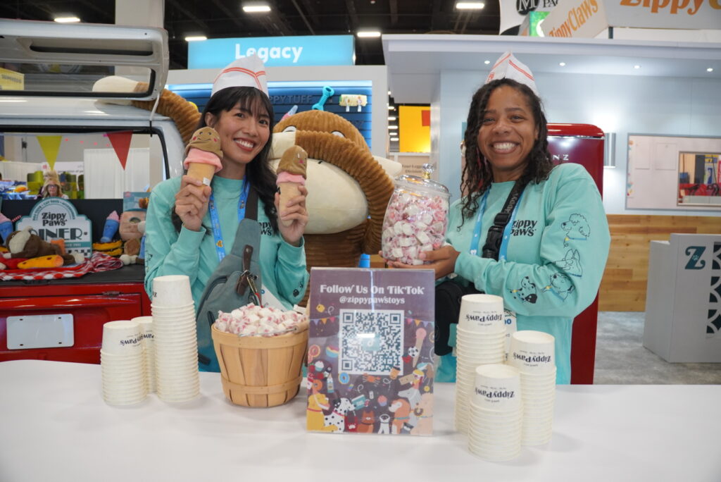 Two women in matching outfits hold ice cream cones and smile behind a counter with cups, a jar of marshmallows, a basket of marshmallows, and a TikTok QR code sign at an event booth.