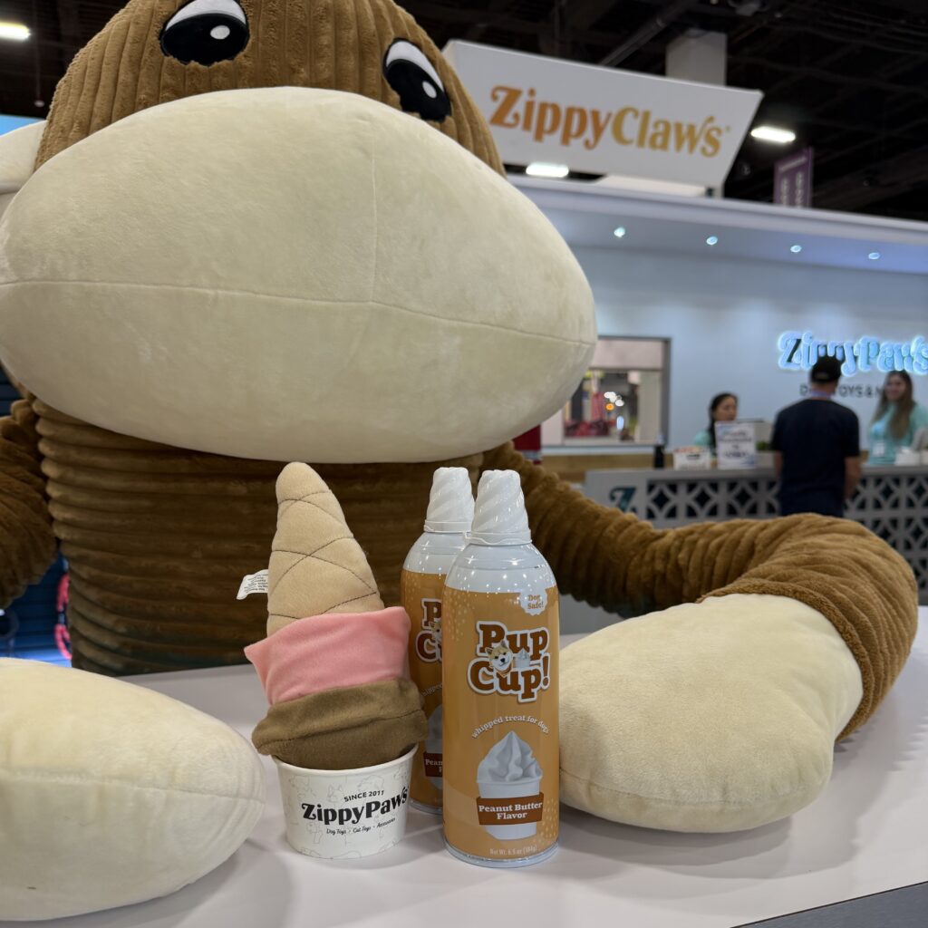 Large stuffed animal sits behind display of ZippyPaws toys and two cans of Pup Cup peanut butter. ZippyPaws booth and people visible in the background.