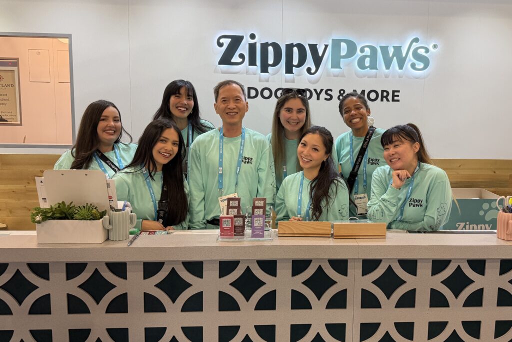 Nine people wearing matching mint-green shirts stand behind a counter at the ZippyPaws booth during SuperZoo 2026, smiling for a group photo.