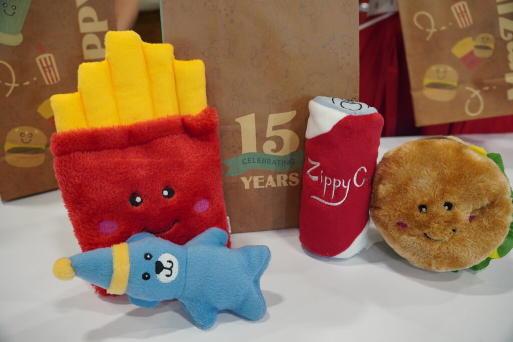 Four plush toys—a smiling fries, a blue star with a hat, a soda can, and a hamburger—are arranged in front of a “15 Years Celebrating” sign on a white surface.