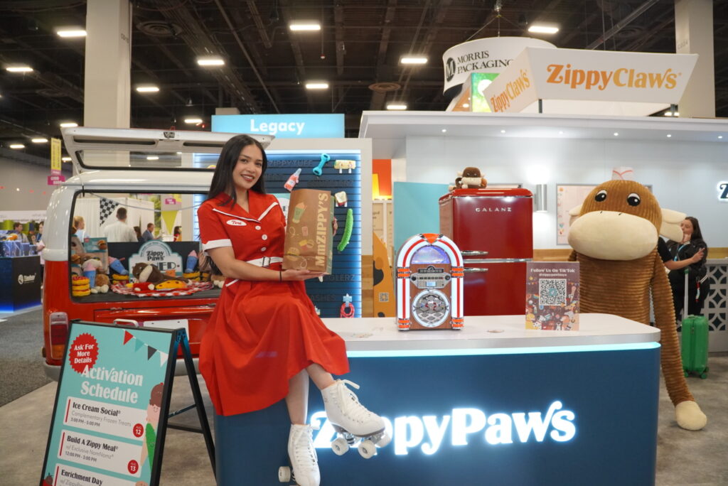 A woman in a red dress sits on a ZippyPaws booth counter, holding a product bag, with colorful displays, a jukebox, and a large plush dog in the background at a trade show.