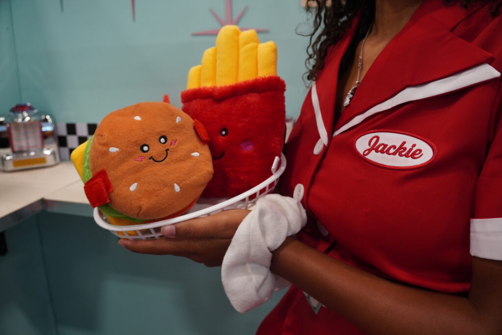 A person in a red diner uniform holds a basket with plush toys of a smiling burger and fries.