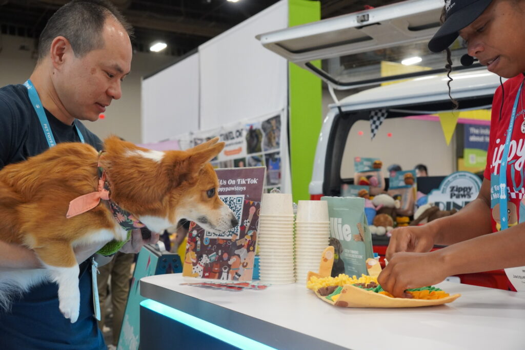 A man holds a corgi while a person prepares a dog-friendly meal at a counter with cups, signs, and a van in the background at an indoor event.