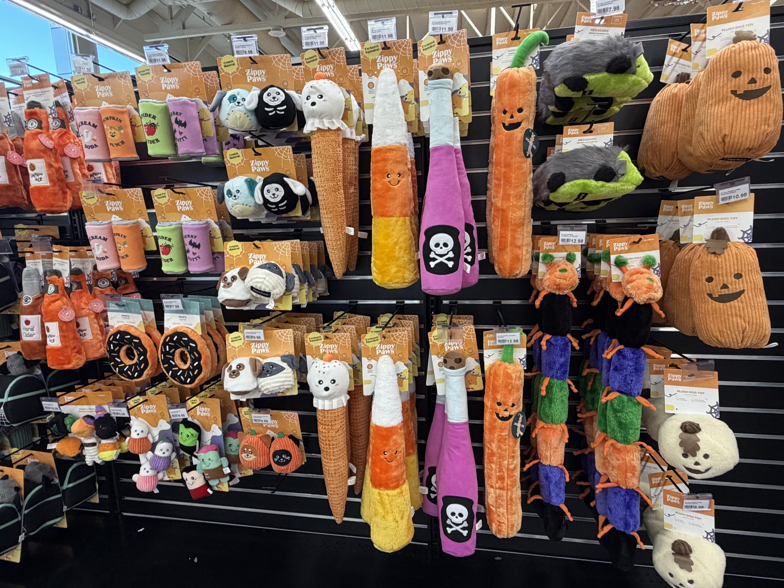 Halloween-themed dog toys including plush pumpkins, carrots, bats, skulls, and ghosts are displayed on black metal racks in a pet store.