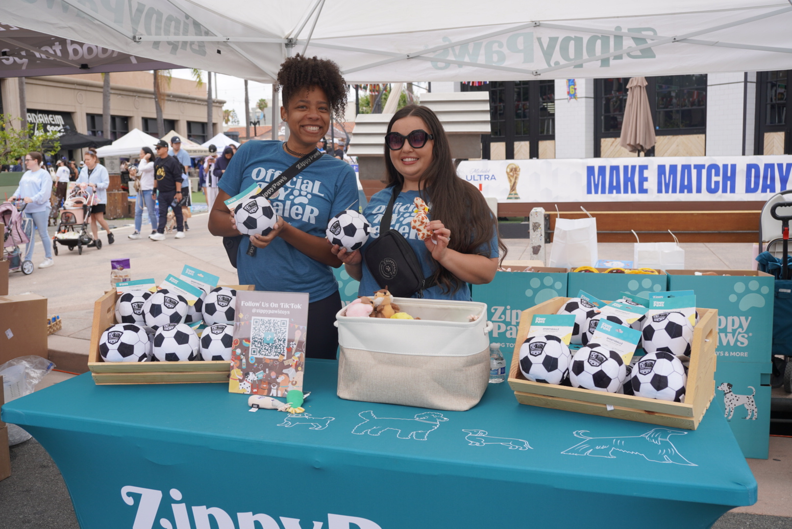 Two women stand behind a ZippyPaws booth displaying soccer ball dog toys at an outdoor event, smiling at the camera.