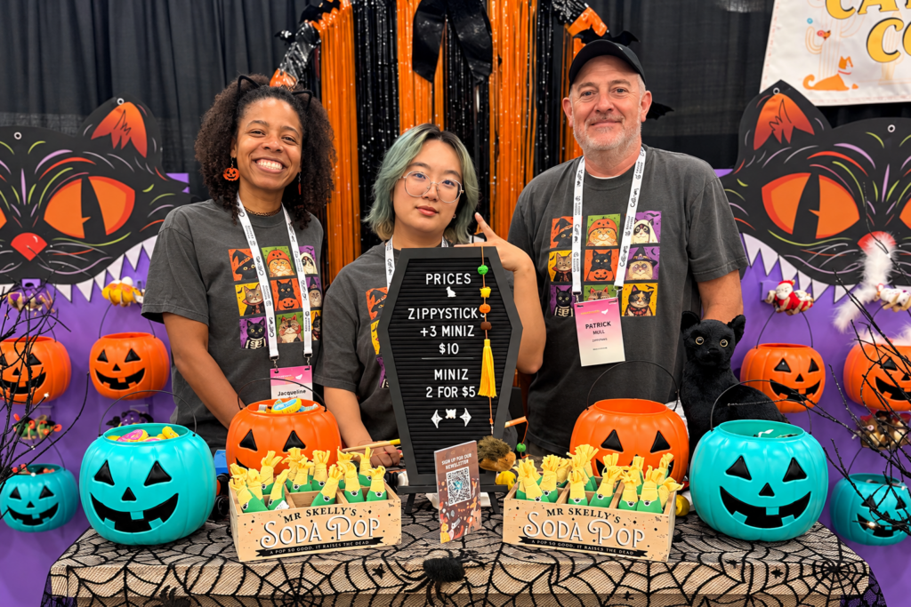 Three people stand behind a table decorated for Halloween, featuring “Mr. Skelly’s Soda Pop” crates, jack-o’-lantern buckets, and a sign listing product prices.