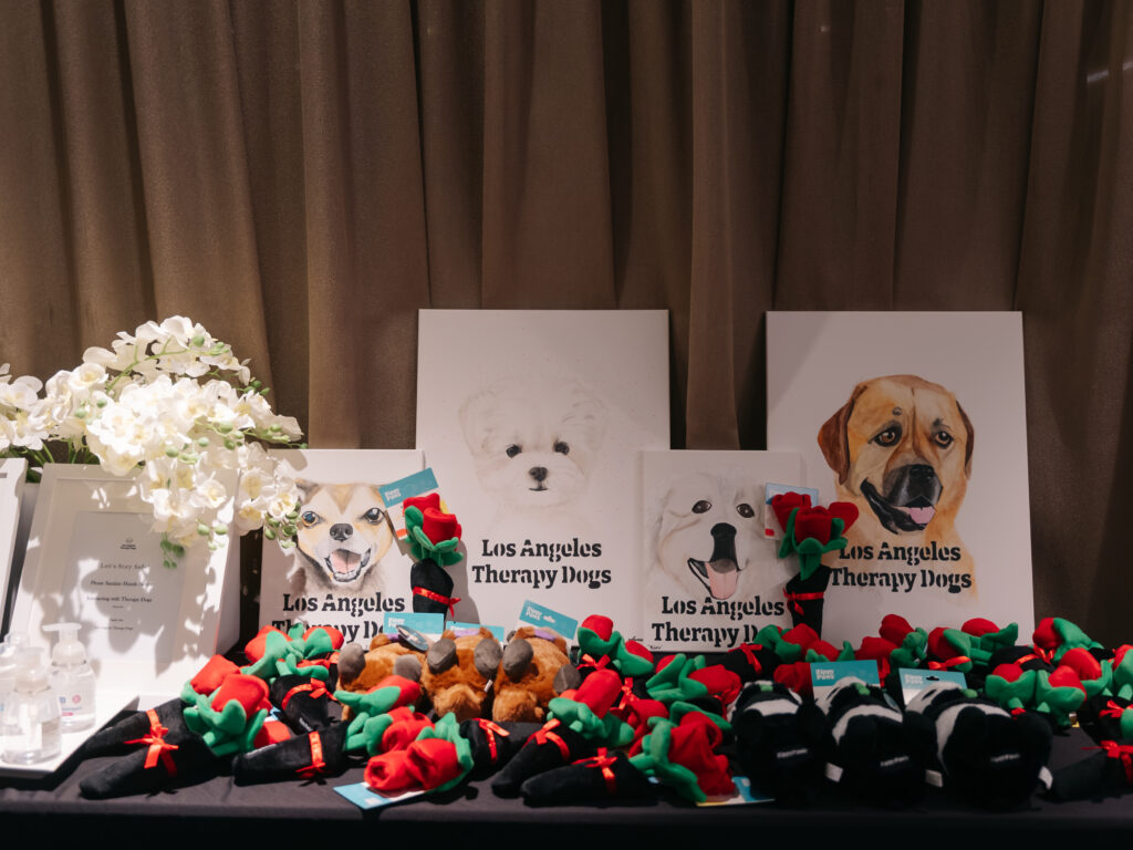 A table display for Los Angeles Therapy Dogs featuring dog portraits, small white flower arrangements, and many plush roses.