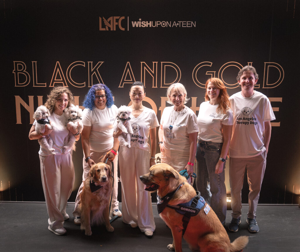 Six volunteers from Los Angeles Therapy Dogs posing for a photo in front of a “Black and Gold” event backdrop, with two dogs being held and two dogs sitting on the floor.