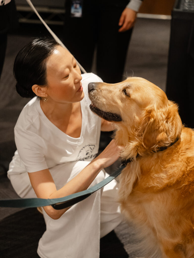 A woman in white clothing kneels to pet a golden retriever on a leash.