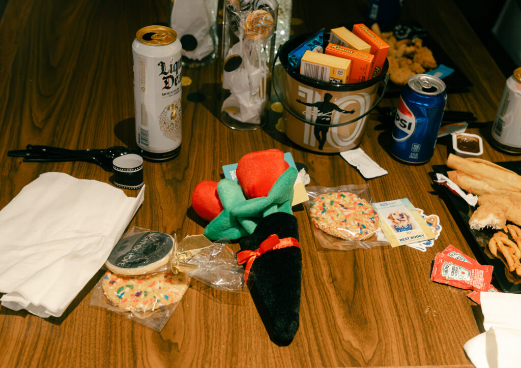 A wooden table scattered with snacks, including cookies, a bucket of candy, a drink can, and a plush carrot toy.