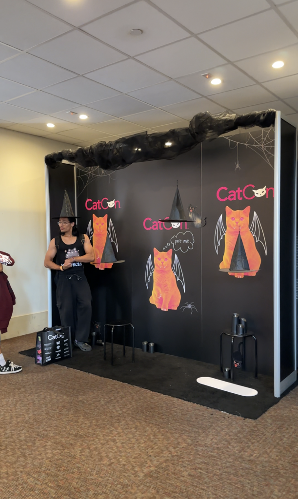 A person in a witch hat stands in a Halloween-themed photo booth featuring illustrations of orange cats with bat wings.
