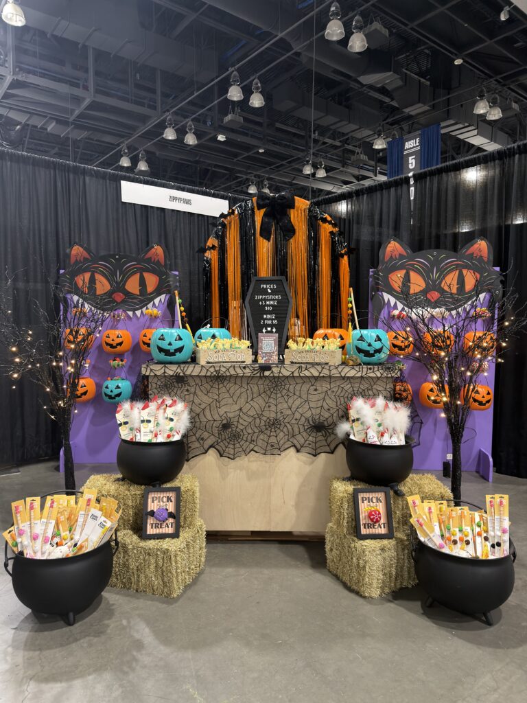 Halloween-themed vendor booth featuring black cat decorations, orange pumpkins, hay bales, and black cauldrons filled with candy, with a price sign for Zippysticks and Miniz.